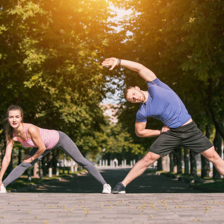 Fit fitness woman and man doing stretching exercises outdoors at park. Couple doing hamstring leg stretching exercise and stretches. Female amd male sports models exercising outdoor in summer.